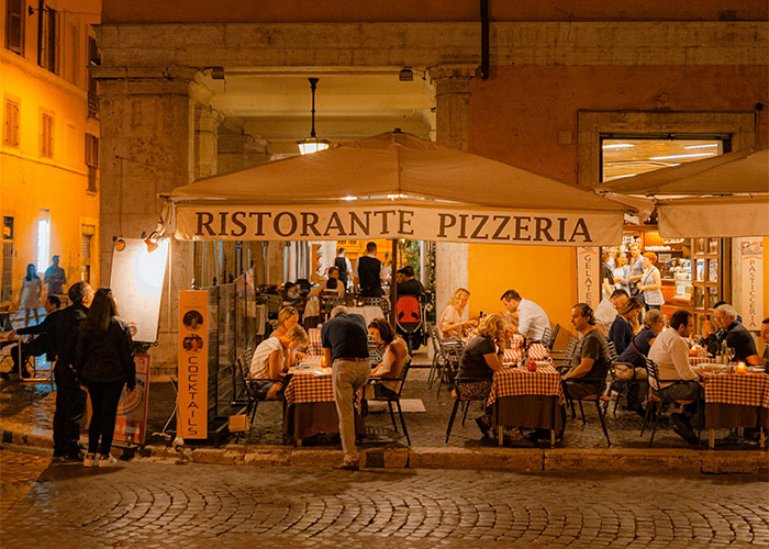 Outdoor Italian pizzeria at night with diners seated under canopy, relating to restaurant owner and tourists sharing pizzas controversy. Outdoor Italian pizzeria at night with diners seated under canopy, relating to restaurant owner and tourists sharing pizzas controversy.