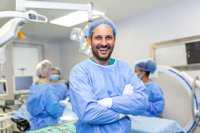 Smiling male surgeon in scrubs and cap stands confidently in operating room during cosmetic surgery mole removal procedure. Smiling male surgeon in scrubs and cap stands confidently in operating room during cosmetic surgery mole removal procedure.