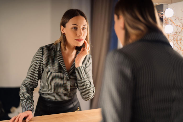 Young woman looking at her face in the mirror, reflecting on plastic surgery and appearance concerns in a dimly lit room. Young woman looking at her face in the mirror, reflecting on plastic surgery and appearance concerns in a dimly lit room.