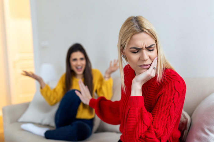 Two sisters in a living room, one looks upset refusing a plastic surgery gift, the other gesturing angrily. Two sisters in a living room, one looks upset refusing a plastic surgery gift, the other gesturing angrily.