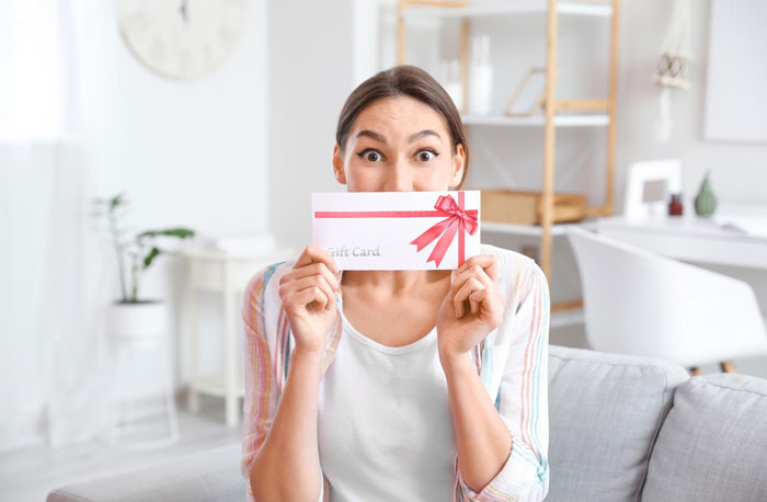 Young woman holding a plastic-surgery gift card with a surprised expression in a bright living room setting Young woman holding a plastic-surgery gift card with a surprised expression in a bright living room setting
