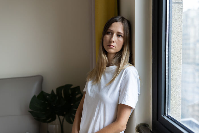 Young woman in a white shirt looking thoughtful by a window, reflecting on feelings only adopted people understand
