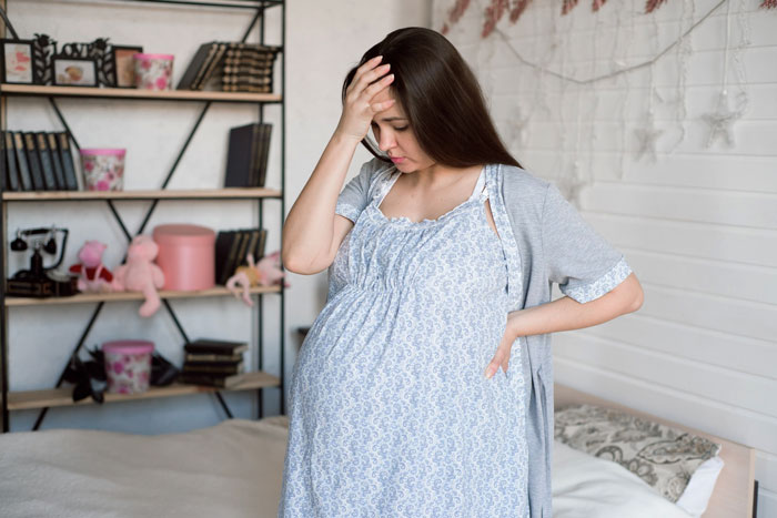Pregnant daughter holding her head, looking worried and standing in a bedroom with shelves and toys in the background.