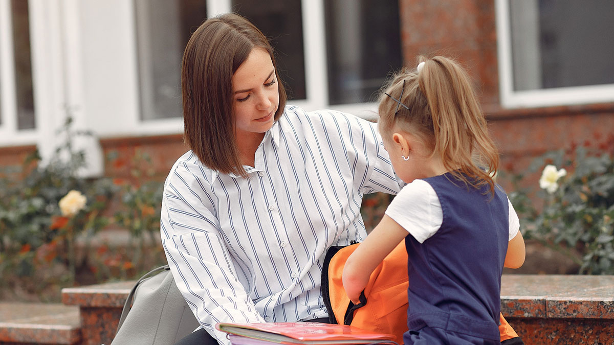 Mother helping her preschool daughter with backpack outside, capturing a moment of parenting at the school entrance.