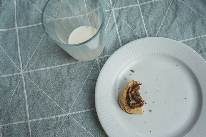 Partially eaten cinnamon roll on a white plate next to a glass with milk on a gray patterned tablecloth, relationship gestures concept.