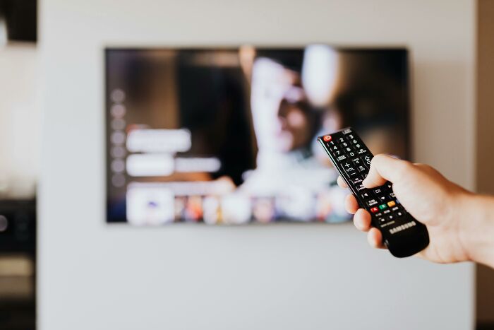 Hand holding a remote control pointing at a blurred TV screen, illustrating relationship gestures and princess treatment.