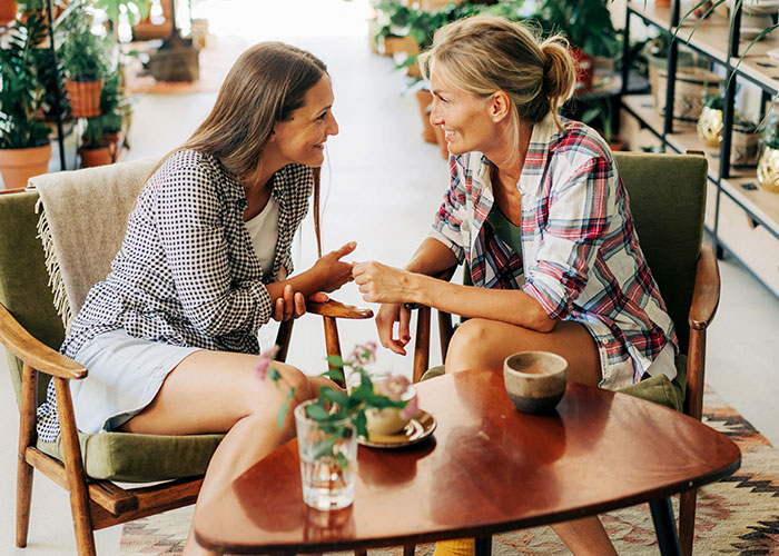 Two women engaged in a friendly conversation using psychological tricks to connect in a cozy, plant-filled café setting.