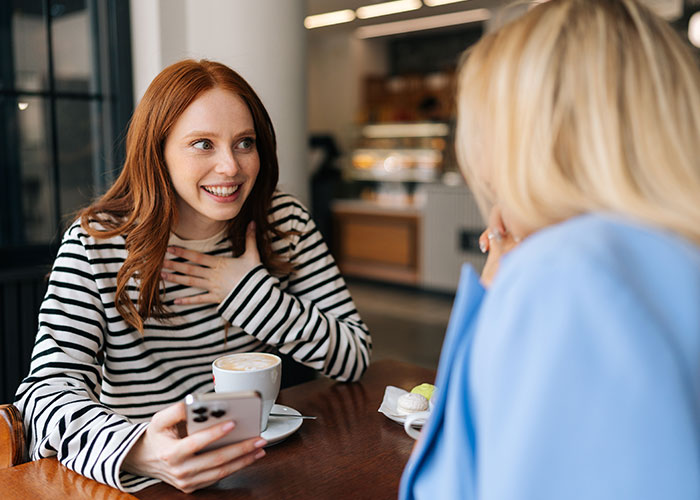 Two women in a cafe using psychological tricks to connect, one smiling and holding a smartphone during a friendly conversation.