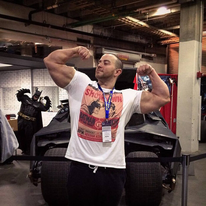 Man flexing muscles in front of a Batmobile with Batman photobombing in the background at a convention.