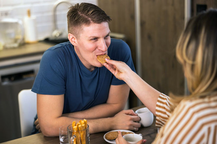 Woman feeding boyfriend a cookie at kitchen table, showcasing perfect secret cookies and a joyful moment together.