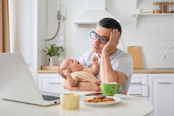 Frustrated man holding baby while working on laptop at home, illustrating babysitting on vacation dilemma. Frustrated man holding baby while working on laptop at home, illustrating babysitting on vacation dilemma.
