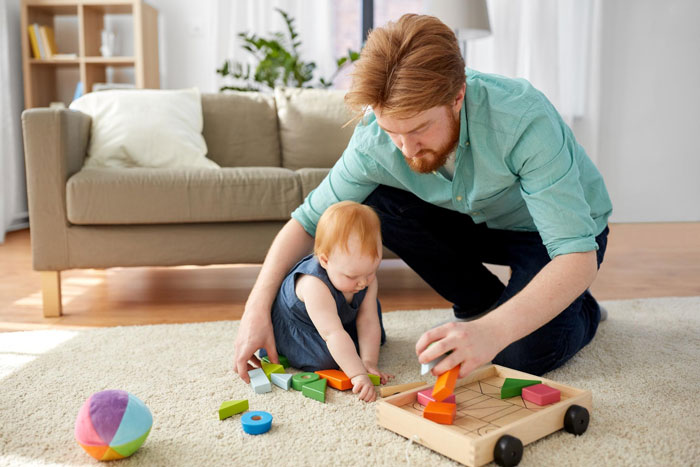 Man spending time with toddler playing with wooden toys indoors, illustrating babysitting on vacation concerns. Man spending time with toddler playing with wooden toys indoors, illustrating babysitting on vacation concerns.