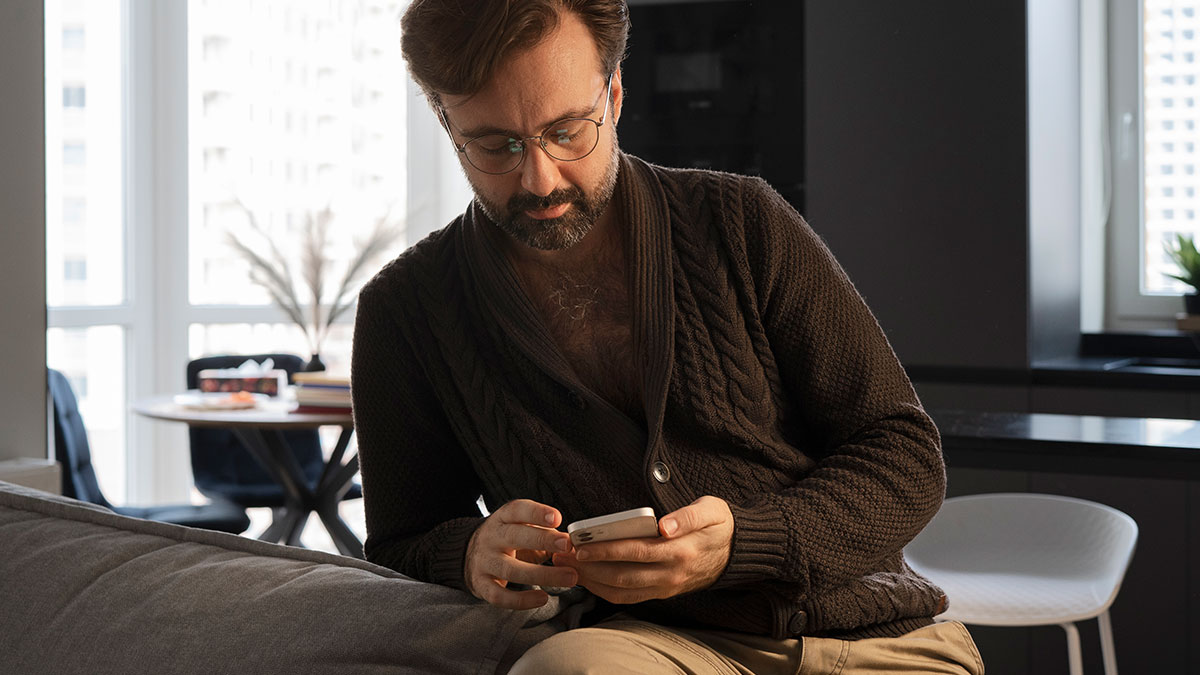 Man in a brown sweater refusing to bring expensive food for Thanksgiving, looking at his phone in a modern living room.