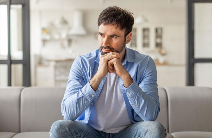 Man in a blue shirt sitting on a couch looking thoughtful and concerned, reflecting on cosmetic surgery recovery help refusal. Man in a blue shirt sitting on a couch looking thoughtful and concerned, reflecting on cosmetic surgery recovery help refusal.