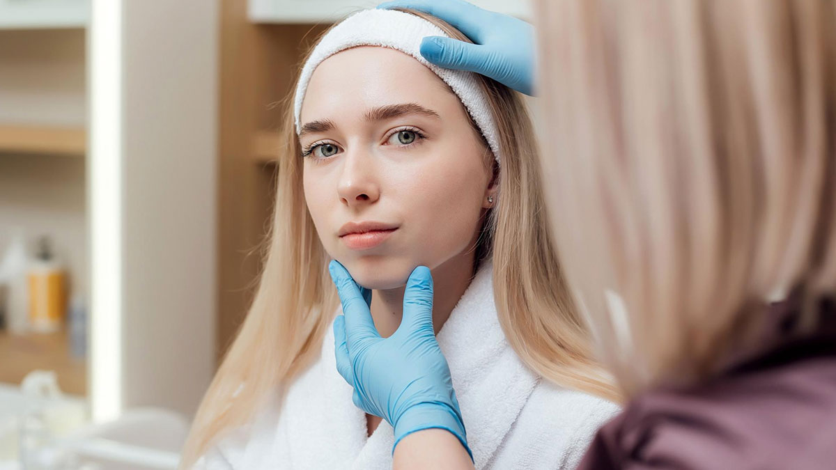 Young woman wearing a headband and white robe, being examined by a professional with blue gloves, related to BBL plans.