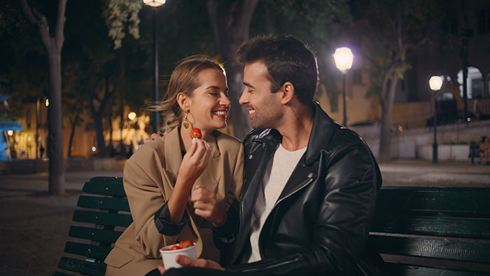 Couple sitting on park bench at night, sharing food and smiling, illustrating how people's lies got way out of hand.