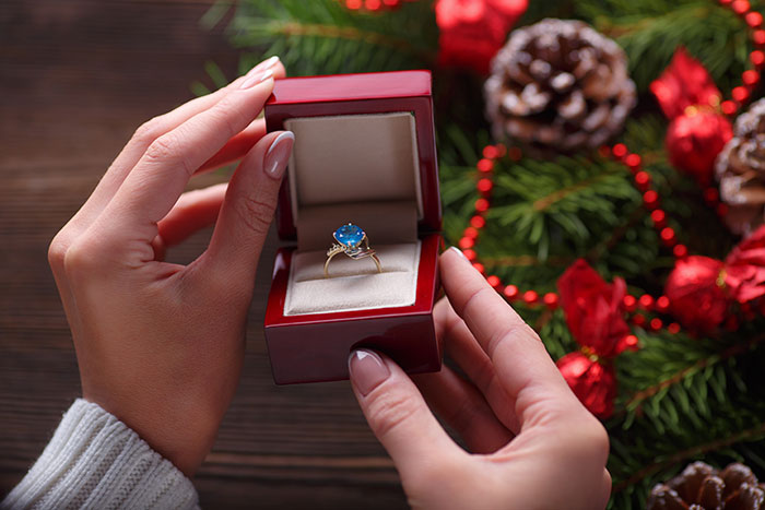 Hands holding a red ring box with a gemstone ring inside surrounded by festive holiday decorations about lies changing lives.