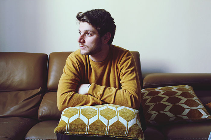 Young man in a mustard sweater sitting on a couch looking away, reflecting on how people's lies got out of hand.