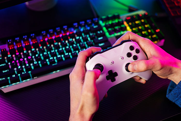 Person holding a white gaming controller, playing video games with colorful RGB keyboard in the background.