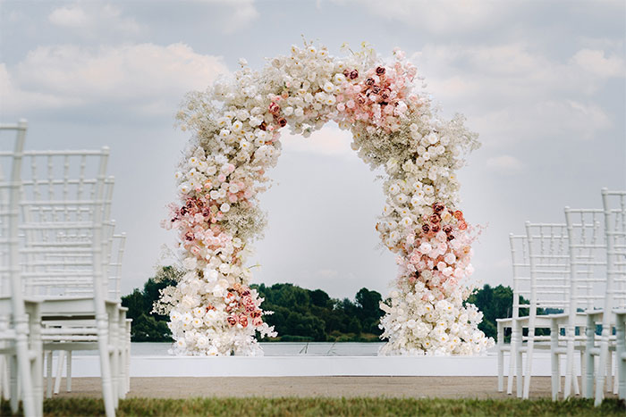 Floral wedding arch setup by a lake with rows of white chairs for a marriage proposal event outdoors.