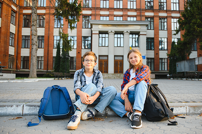 Two stepchildren sitting outside a boarding school with backpacks, waiting for a woman’s request. Two stepchildren sitting outside a boarding school with backpacks, waiting for a woman’s request.