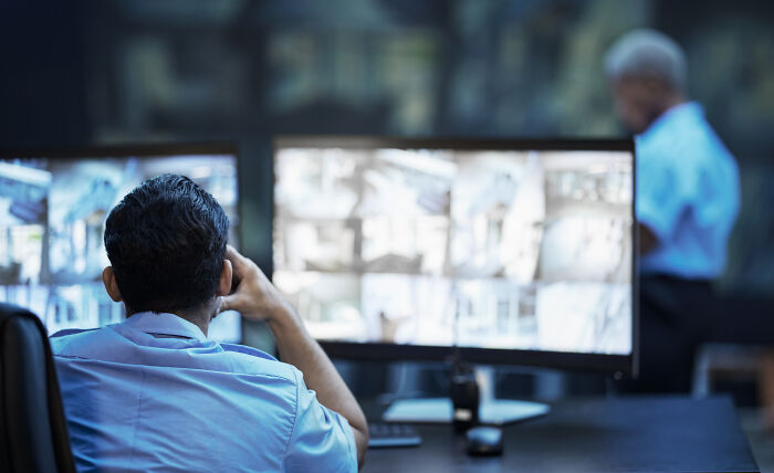 Security personnel monitoring multiple screens displaying surveillance camera footage in a control room environment.