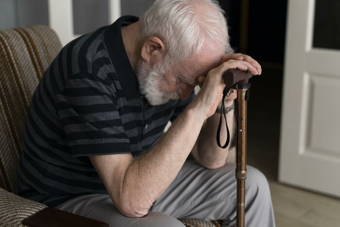 Elderly man sitting with head down, holding a cane, reflecting on regrets about having kids in a quiet room.