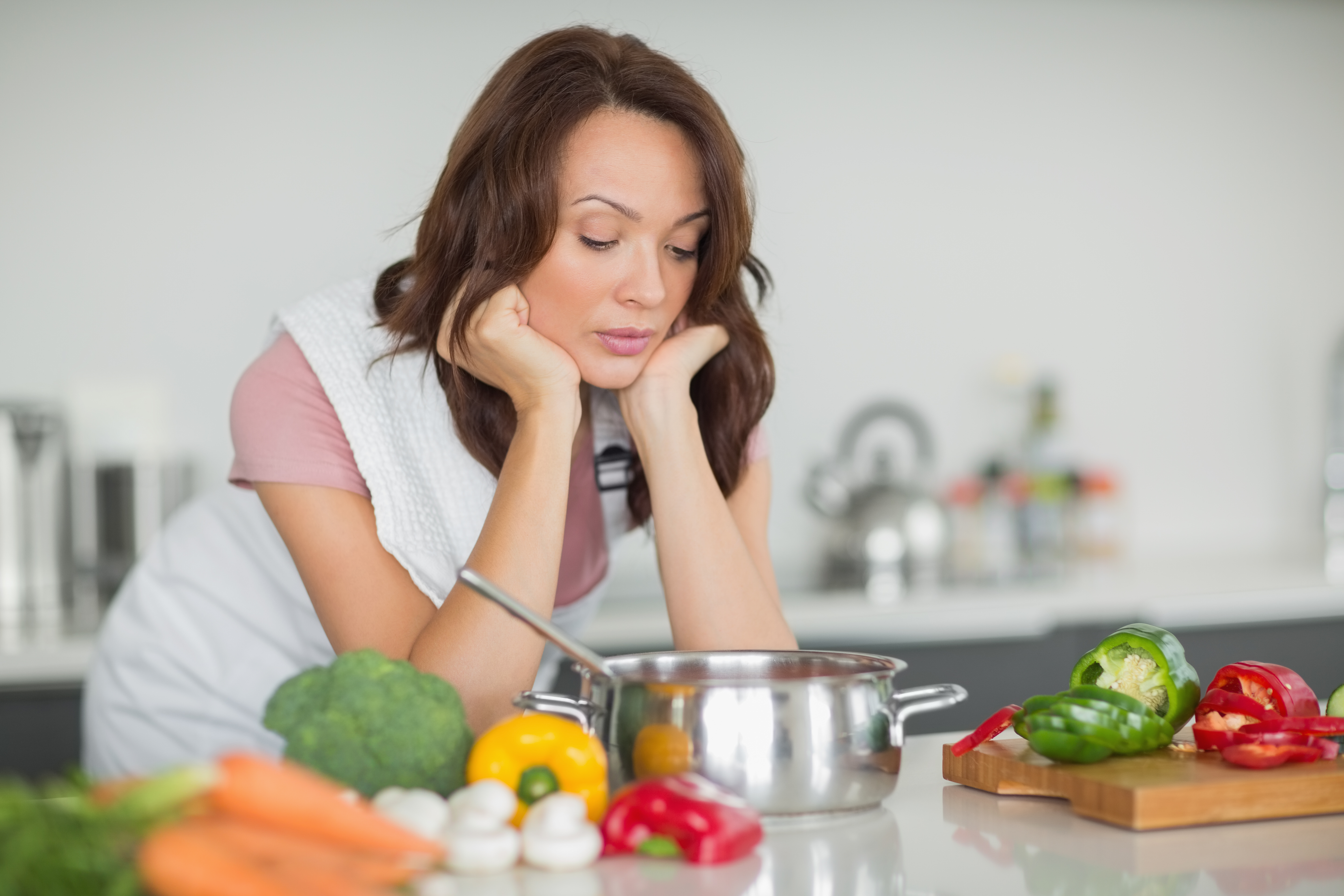 Woman in kitchen looking frustrated while cooking with fresh vegetables and a pot on the counter related to man expecting wife to cook.