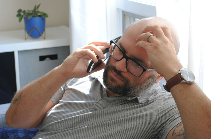 Man with glasses, beard, and watch looking concerned while talking on phone in a bright room with plant and window. Man with glasses, beard, and watch looking concerned while talking on phone in a bright room with plant and window.