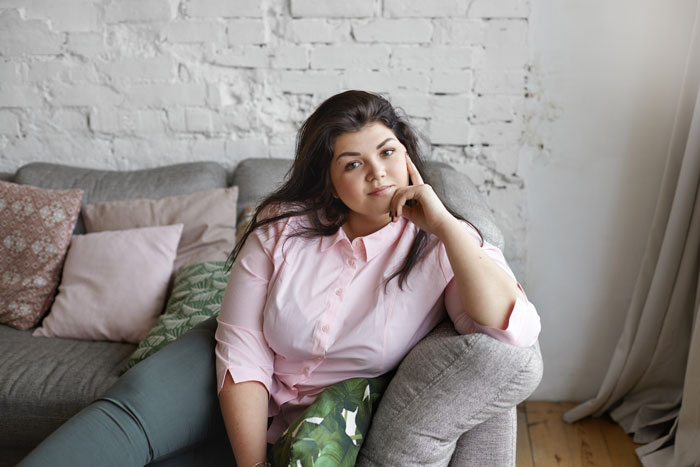 Young woman in pink shirt sitting on couch, appearing thoughtful about refusing to babysit badly behaved nephew. Young woman in pink shirt sitting on couch, appearing thoughtful about refusing to babysit badly behaved nephew.