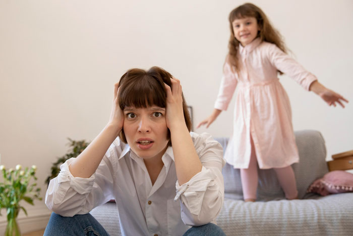 Woman with concerned expression holding her head while her sister plays in a gentle parenting house setting. Woman with concerned expression holding her head while her sister plays in a gentle parenting house setting.