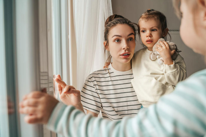 Young sister practicing gentle parenting at home, holding toddler and calmly talking near window in cozy house. Young sister practicing gentle parenting at home, holding toddler and calmly talking near window in cozy house.