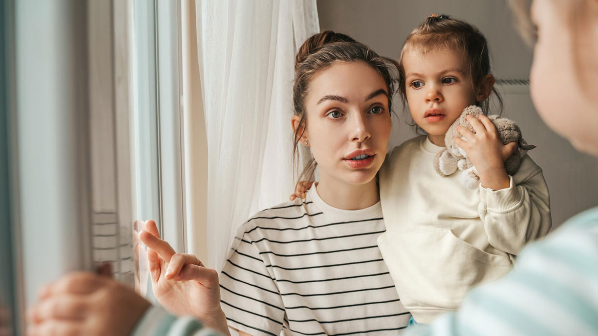 Sister practicing gentle parenting at home, holding child near window while speaking softly in a calm house setting.