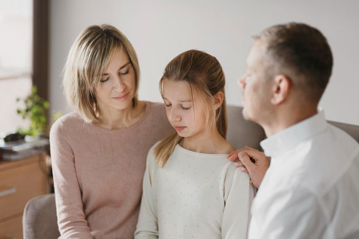 Teen girl looking upset while parents comfort her, highlighting struggles with mentally unstable sister and family priorities.