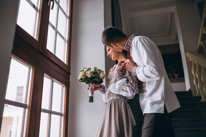 Couple in traditional clothing holding flowers by a large window, representing a big wedding scene. Couple in traditional clothing holding flowers by a large window, representing a big wedding scene.