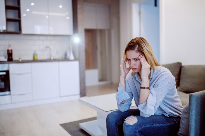 Young woman sitting in living room looking stressed and upset, illustrating rejection sensitive dysphoria in family conflict. Young woman sitting in living room looking stressed and upset, illustrating rejection sensitive dysphoria in family conflict.