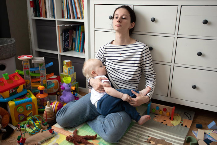 Woman sitting on floor holding baby in a cluttered room, expressing concern after spotting roaches following sister's move-in. Woman sitting on floor holding baby in a cluttered room, expressing concern after spotting roaches following sister's move-in.