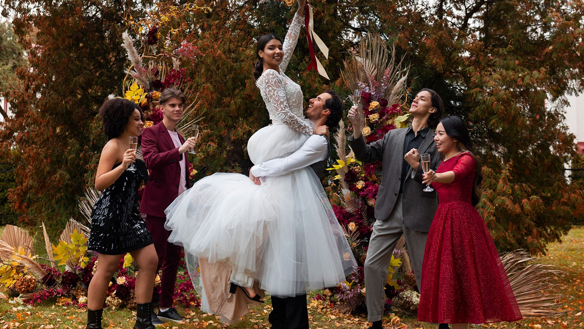 Bride lifted by groom with friends celebrating outdoors in wedding money culture setting with autumn decorations and toasts.