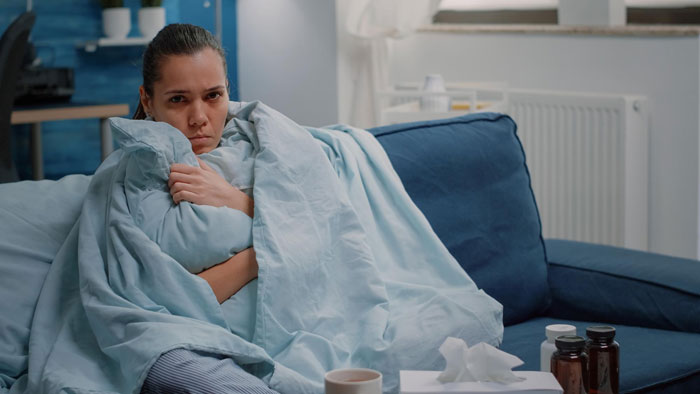 Sick woman wrapped in blanket on couch, with medicine bottles and tissues nearby, showing signs of illness and discomfort. Sick woman wrapped in blanket on couch, with medicine bottles and tissues nearby, showing signs of illness and discomfort.