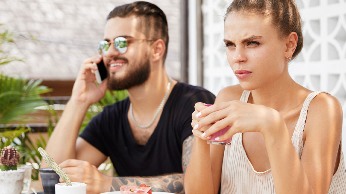 Man showing affection to his sick wife while sister looks annoyed during a tense outdoor cafe moment.