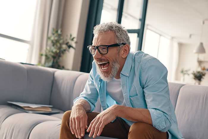 Man laughing joyfully while sitting on a couch in a bright living room after teen breaks new chair