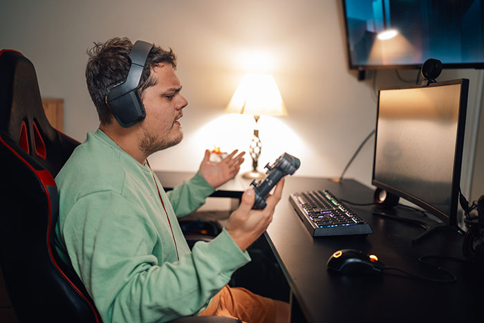Teen gamer frustrated sitting on broken chair, wearing headset and holding controller in front of computer setup at home. Teen gamer frustrated sitting on broken chair, wearing headset and holding controller in front of computer setup at home.