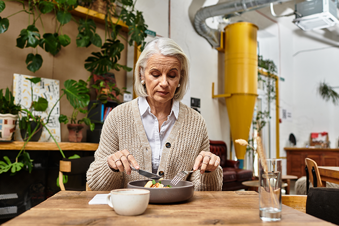 Older woman sitting at a table with a plate of food, appearing thoughtful in a cozy, plant-filled dining space.