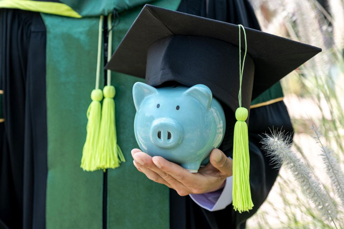 Person in graduation gown holding piggy bank with graduation cap, symbolizing college fund and savings. Person in graduation gown holding piggy bank with graduation cap, symbolizing college fund and savings.