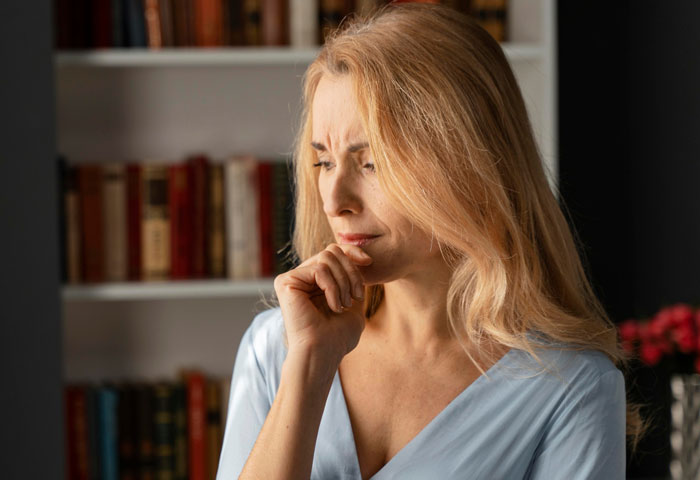 Pensive woman with long blonde hair in a blue blouse thinking at home with a bookshelf in the background. Pensive woman with long blonde hair in a blue blouse thinking at home with a bookshelf in the background.