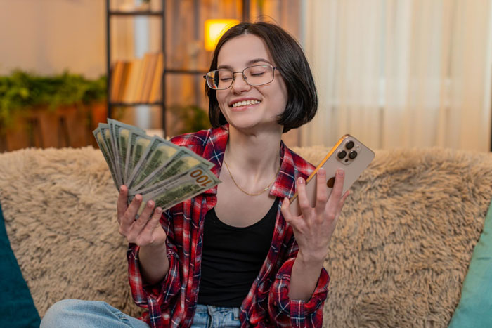 Young woman smiling and holding cash and smartphone indoors, illustrating pregnant SIL paying nieces for help scenario.