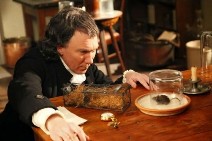 Man in historical clothing leaning over a table with a mouse in a glass jar, illustrating strange facts about our world.