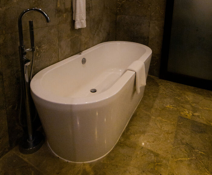 White modern bathtub in a hotel bathroom with marble floors and walls, towel draped over the edge of the tub. White modern bathtub in a hotel bathroom with marble floors and walls, towel draped over the edge of the tub.