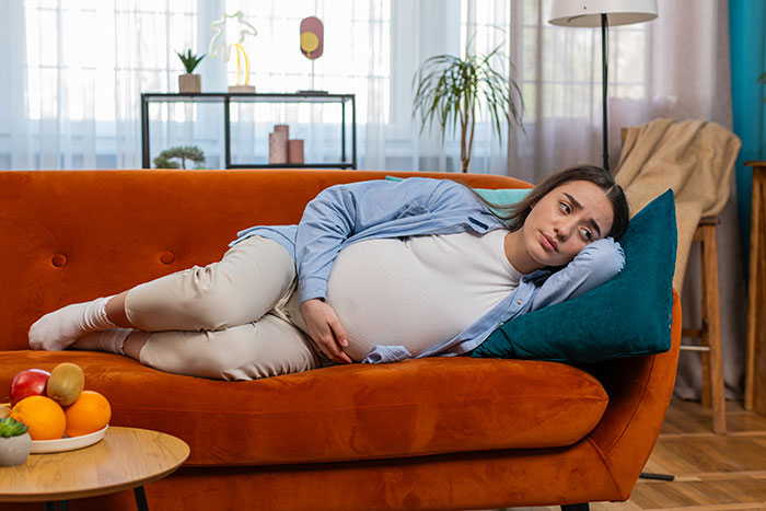 Pregnant woman lying on an orange couch looking upset, illustrating tension with a golden child sibling about meals. Pregnant woman lying on an orange couch looking upset, illustrating tension with a golden child sibling about meals.