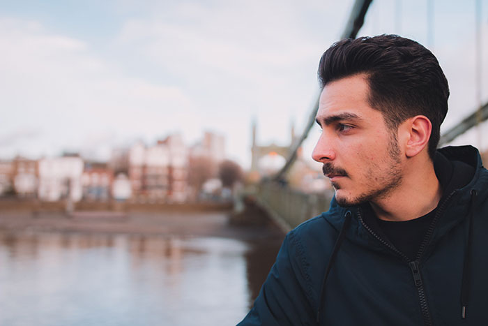 Young man in a dark jacket looking thoughtfully across a river with a blurred cityscape in the background, revealing emotions.